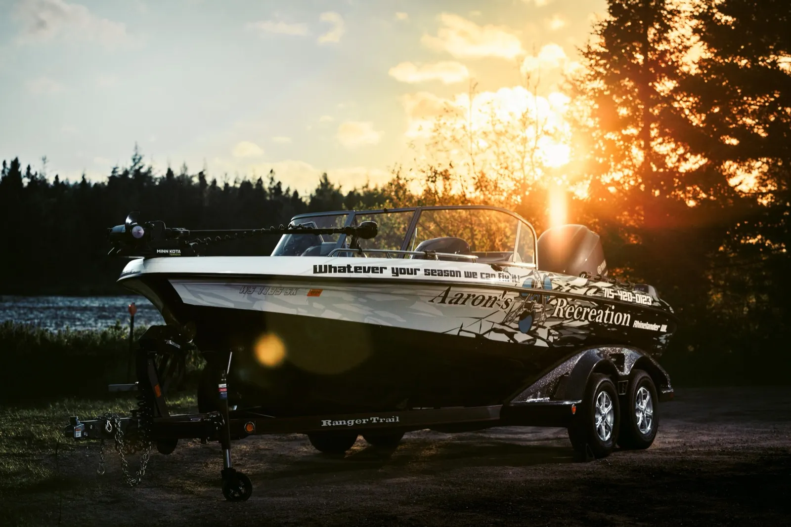 Aaron's Recreation boat and trailer beside the lake, with Whatever your season we can fix it painted on the hull